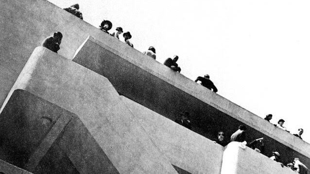 Image of a brutalist building with many balconies and staircases, with people looking over the edge