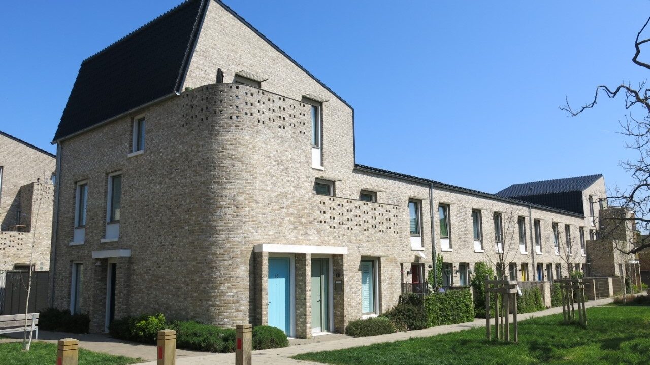 Photograph of a grey building against a blue sky. There are bollards to the left of the building and grass and trees in front of it.
