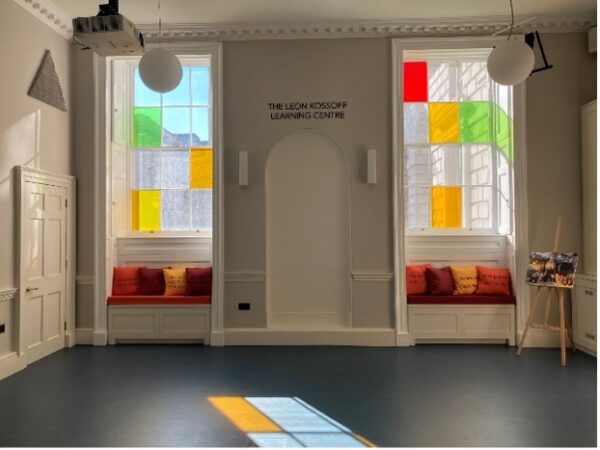 Still of the Leon Kossoff Learning Centre with no tables or chairs. Light streams in from two large windows in the ornate but friendly room; the windows have three and four scattered panes respectively (out of twelve total on each window) that have brightly coloured glass in them.