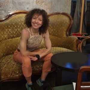 Lisa is seated and leaning forward with a welcoming smile and has large, curly hair.
