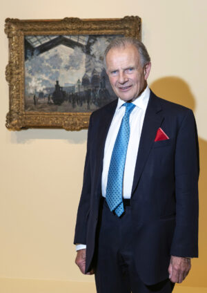 Christopher Mclaren with a suit and blue tie standing in front of an impressionist painting with a gilt frame